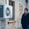 Un homme vêtu d'un manteau d'hiver regarde une pompe à chaleur installée sur le mur fissuré d'une vieille maison.